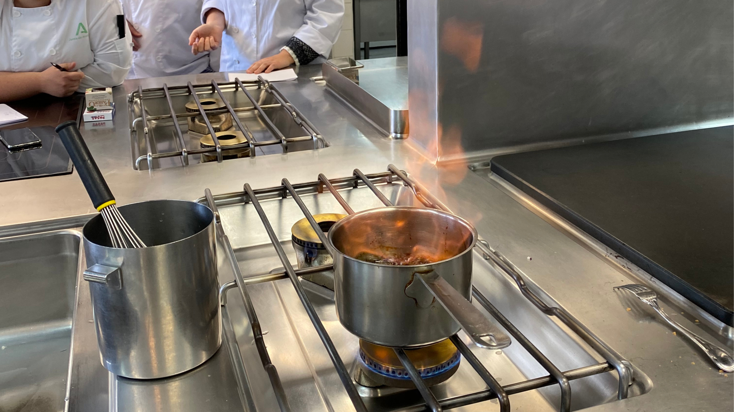 Cocineros con uniforme blanco trabajando en la cocina industrial de un comedor colectivo, con un cazo al fuego