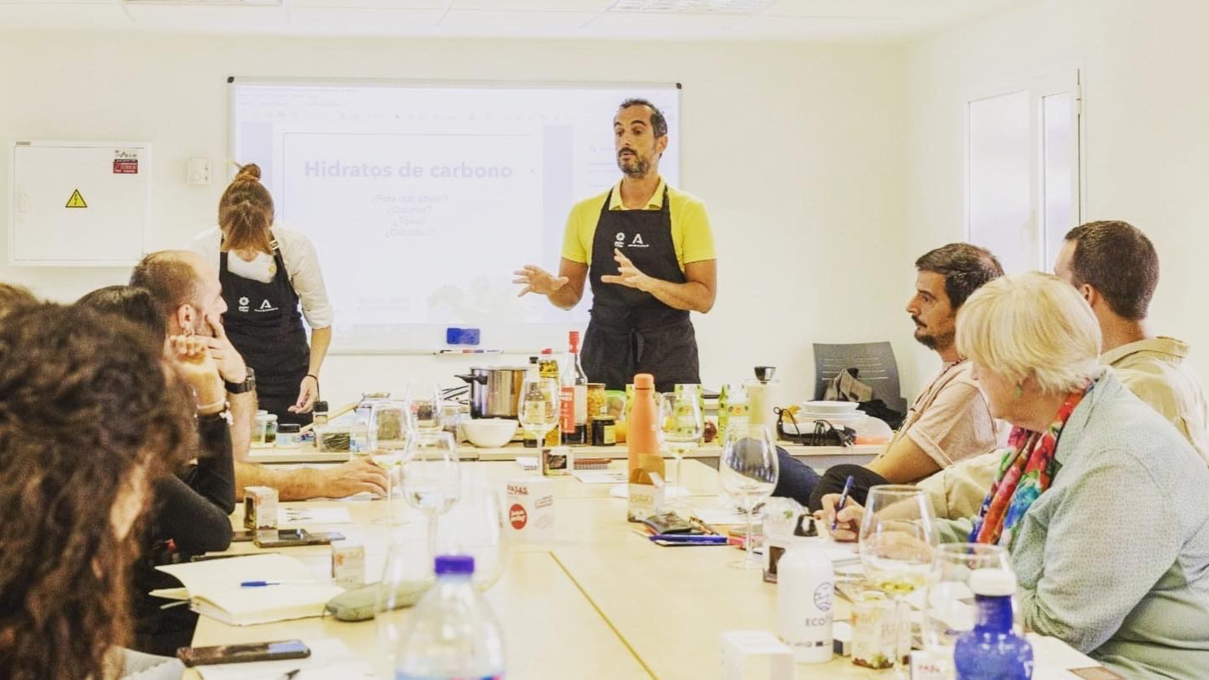 Formador de La Habitación Saludable, con delantal, explicando hidratos de carbono a empleados en una sala de reuniones